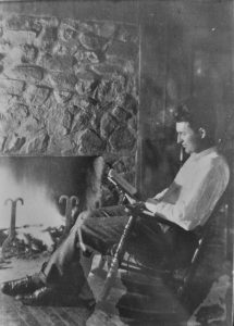 Black and white photo of Paul Green sitting in a rocking chair by a stone fireplace, reading a book