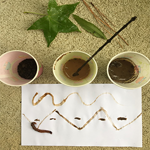 Three cups with different colors of paint made from soil, and a paper with designs made with the soil paint.