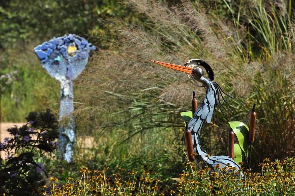 A metal great blue heron stands in front of an abstract blue cast iron sculpture, surrounded by black-eyed susans and switchgrass.