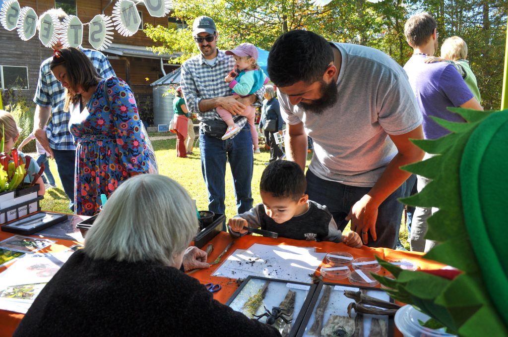 A boy examines soil with a magnifying glass at one of the NatureFest activity tables. His father stands behind him. More parents and children are in the background.