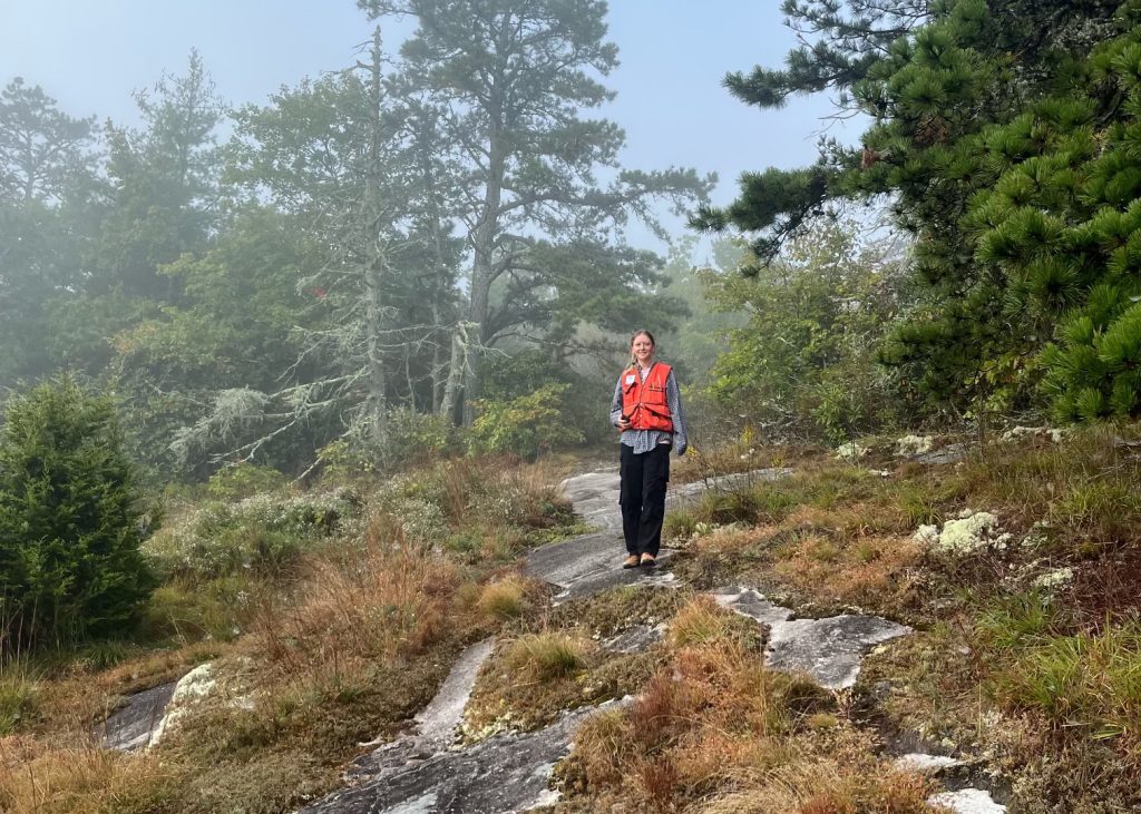 Sophie stands on a rocky, misty mountainside wearing a red vest for seed collection